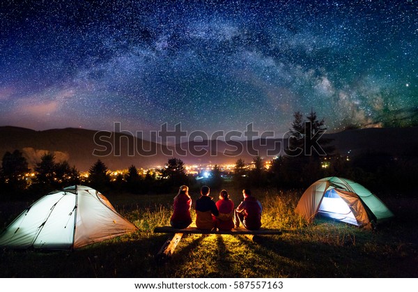 Friends hikers sitting on a bench made of logs and\
watching fire together beside camp and tents in the night. On the\
background beautiful starry sky, mountains and luminous town. Rear\
view
