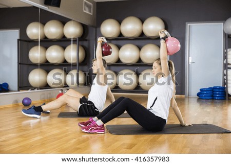 Friends Lifting Kettlebells While Sitting On Mat In Gym