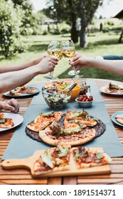 Friends Making Toast During Summer Picnic Outdoor Dinner In A Home Garden. Close Up Of People Holding Wine Glasses With White Wine Over Table With Pizza, Salads And Fruits. Dinner In A Orchard