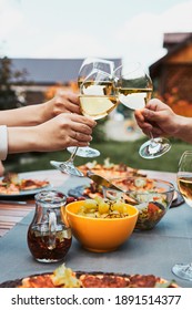 Friends Making Toast During Summer Picnic Outdoor Dinner In A Home Garden. Close Up Of People Holding Wine Glasses With White Wine Over Table With Pizza, Salads And Fruits. Dinner In A Orchard