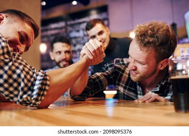 Friends plays arm wrestling. Group of people together indoors in the pub have fun at weekend time.