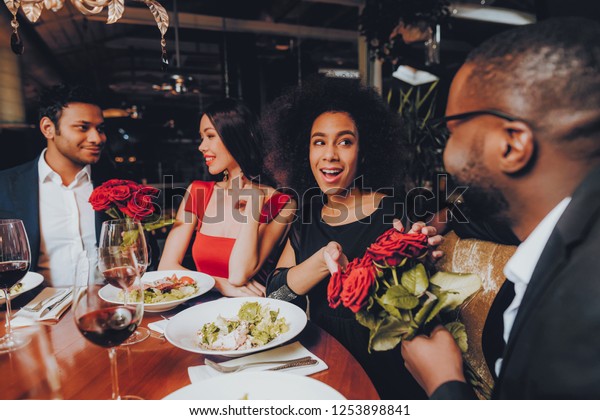 Friends in a Restaurant Enjoying Romantic\
Evening. Group Happy Friends Enjoying Dating in Restaurant. Double\
Couple Dating in Restaurant. Romantic Concept. Bouquet Red Roses.\
Love Concept.