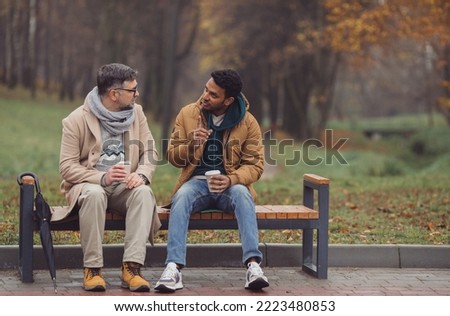 Friends, a senior and a young man sit in the park on a bench and talk in the autumn park.