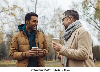Friends, a senior and a young man walking and talking and drinking coffee together in the autumn park. - Shutterstock ID 2223478821
