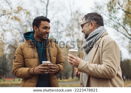 Friends, a senior and a young man walking and talking and drinking coffee together in the autumn park.