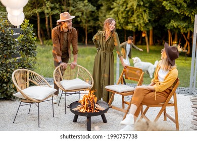 Friends sitting by a fireplace, paying with dog, having great summertime at backyard near the forest. Barbecue in close company in nature