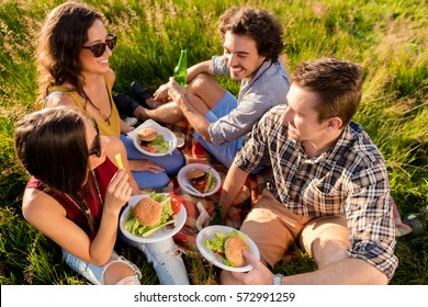 Friends Sitting In Grass And Having Sandwiches At BBQ Party In Meadow