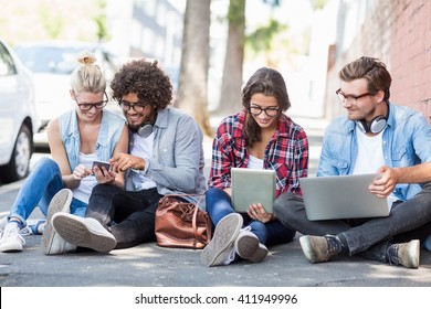 Friends Sitting On Roadside Using Mobile Phone, Digital Tablet And Laptop