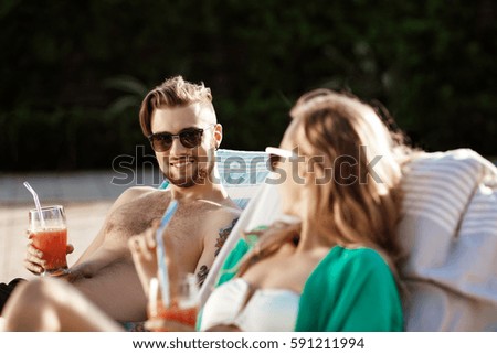 Friends smiling, drinking cocktails, lying on chaises near swimming pool.