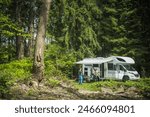 Friends standing in front of a parked camper van, looking at each other. The camper is parked by a serene lake, with trees in the background.