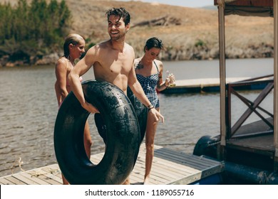 Friends Standing On Floating Dock After Swimming In The Lake. Man Standing Near A Lake With An Inflatable Rubber Tube With His Female Friends Drinking Beer.