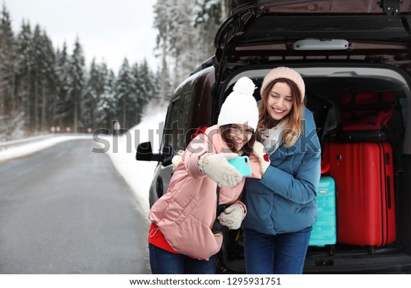 Friends taking selfie near open\
car trunk full of luggage on road, space for text. Winter\
vacation