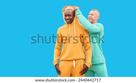  friends. Two men different color black African-American ethnicity and white Caucasian ethnicity standing isolated blue background. One man put on hood to other male Foto stock © 