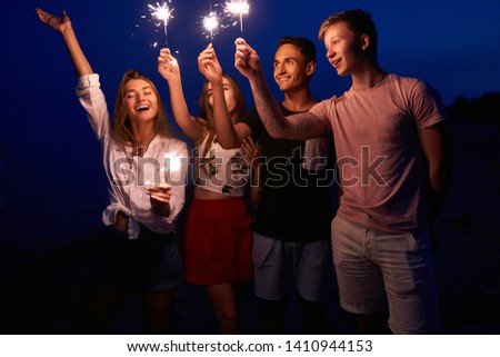 Friends walking, dancing and having fun during night party at the seaside with bengal sparkler lights in their hands. Young teenagers partying on the beach with fireworks. Slow motion steadycam shot.