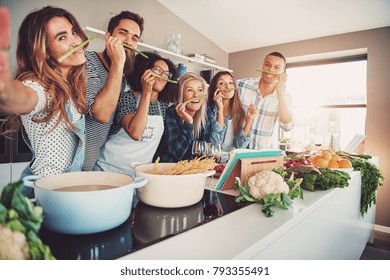 Friends wearing asparagus stalks under their noses while standing in front of table full of pots, pasta noodles and vegetables - Powered by Shutterstock