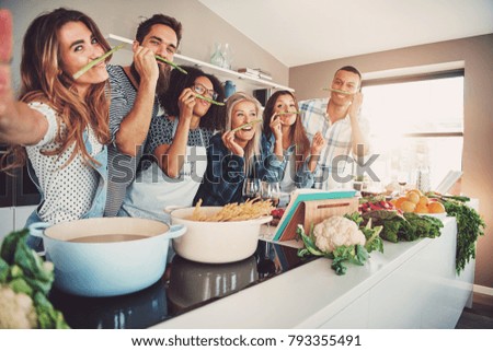 Friends wearing asparagus stalks under their noses while standing in front of table full of pots, pasta noodles and vegetables