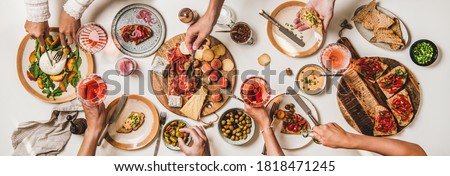 Friends wine and snacks party. Flay-lay of peoples hands with rose wine and food over table with cheese, fruit, smoked meat, tomato brushettas, buratta salad, top view. Wine tasting, gathering concept