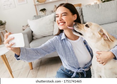 Friendship Concept. Excited beautiful caucasian woman taking selfie with her dog at home, puppy licking her face