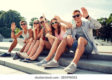 Friendship, Leisure, Summer, Gesture And People Concept - Group Of Smiling Friends Sitting On City Street And Waving Hands