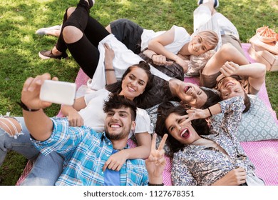 friendship, leisure and technology concept - group of happy smiling friends taking selfie by smartphone chilling on picnic blanket at summer park