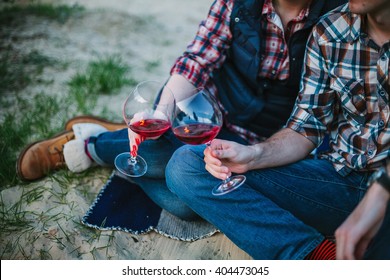 Friendship, Love, Happiness, Summer Vacation, Holidays And People Concept - Couple Sitting On Beach And Drinking Wine From Glasses Near Bonfire In The Evening. Shallow Focus.