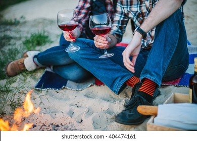 Friendship, Love, Happiness, Summer Vacation, Holidays And People Concept - Couple Sitting On Beach And Drinking Wine From Glasses Near Bonfire In The Evening. Shallow Focus.