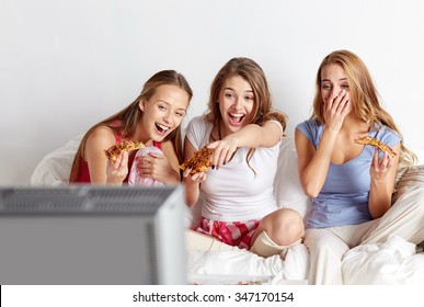 friendship, people, pajama party, entertainment and junk food concept - happy friends or teenage girls eating pizza and watching movie or tv series at home - Powered by Shutterstock