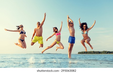 Friendship, Sea, Summer Vacation, Holidays And People Concept - Group Of Smiling Friends Wearing Swimwear And Sunglasses Jumping On Beach