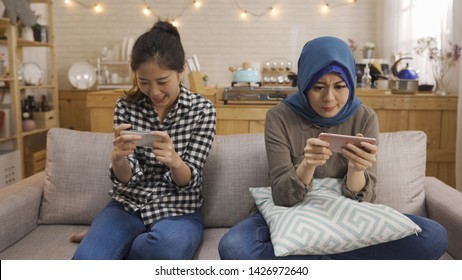 Friendship Technology Home Concept. Two Smiling Multiracial Female Friends With Smartphones In Kitchen. Asian Chinese And Muslim Women Having Fun Playing Video Game Mobile Phone Sitting On Couch Sofa