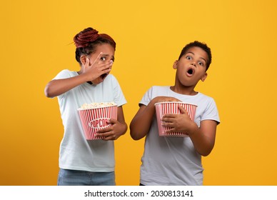 Frightened afro-american boy and girl siblings or friends with popcorn buskets watching horror, sister closing her face, bother grimacing, children watching spooky movie together, yellow background
