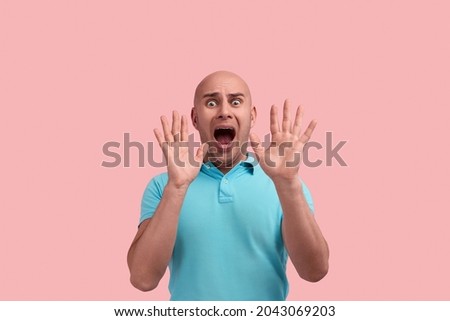 Frightened bald homosexual man with bristle, screams in fright, hold hands near face in protective gesture, has scared expression, gay friendly, wears blue polo shirt, poses over pink background