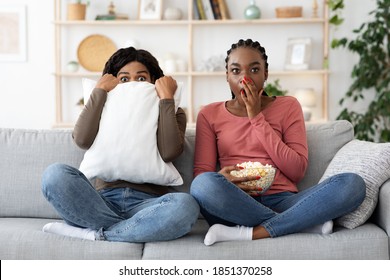 Frightened black girlfriends watching horror together at home, eating popcorn. Scared african american women sitting on sofa, hiding behind pillow and covering mouth with palm, watching movie