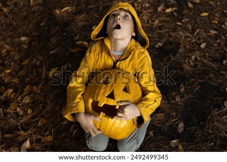 frightened child in an orange jacket sits on the ground strewn with fallen leaves, holding a large Halloween pumpkin in his hands. Evening forest. mood to scare and have fun. Trick or treat