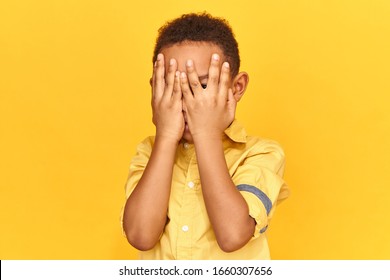 Frightened Dark Skinned Little Boy Covering Face With Both Hands Being Afraid Of Watching Scary Movie, Peeping Through Hole Between Fingers. Confused Timid Child Feeling Shy Hiding His Emotions