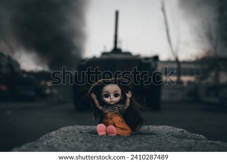A frightened doll sits on the ground surrounded by fire and smoke. A child's toy on the background of an attacking tank. The concept of the horrors of military conflicts. War and children concept.