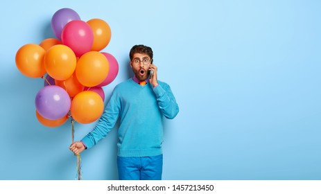 Frightened Embarrassed Man Talks Via Mobile Phone, Finds Out Party Is Postponed, Holds Air Balloons, Wears Blue Jumper And Pants, Stands Indoor. Attractive Male Organizes Party. Monochrome Shot