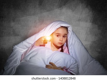 Frightened Girl With Flashlight Hiding Under Blanket. Scared Kid Lying In His Bed At Home. Night Terrors Of Child. Little Girl Afraid Of Dark. Child In Pajamas On Background Of Grey Wall.