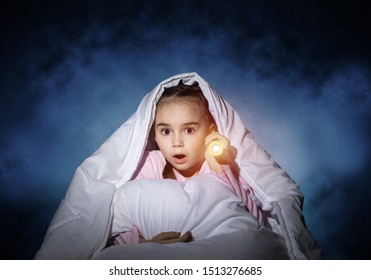 Frightened Girl With Flashlight Hiding Under Blanket. Scared Kid Lying In His Bed At Home. Night Terrors Of Child. Little Girl Can Not Sleep At Night. Child In Pajamas On Background Of Deep Blue Sky