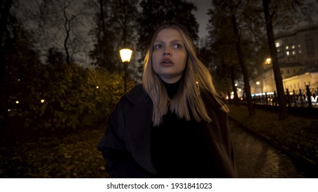 Frightened Girl Running Alone In The Park At Night. Action. Blond Young Girl Is Afraid Of Someone Following Her, Looking Around.