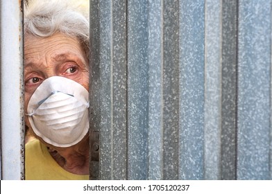 A frightened grandmother in a mask peeps out of the gap in the fence through the gate of her house. Isolation. Coronavirus covid-19. Quarantine elderly people. Stay home. Agoraphobia, mental disorder