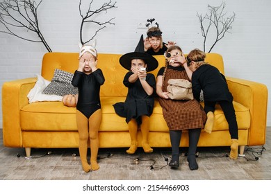 Frightened Kids Dressed For Halloween Sitting On A Yellow Couch, Watching Scary Moment In A Movie. Covering Their Mouths And Eyes.