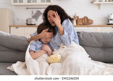 Frightened Mom And Little Kid Son With Bowl Of Popcorn Watching Scary Scene In Movie Closing Their Eyes, Sitting On Sofa At Home. Mother Hugs The Child, Watches A Horror Movie, Feels Fear.