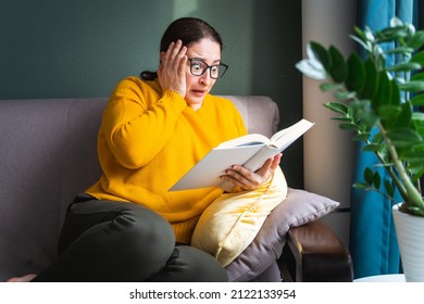 Frightened scared middle-aged woman in eyeglasses reading horror novel, thriller or scary stories while sitting on sofa at home, emotional shocked female with book in hands feeling terrified