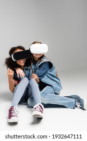 Frightened And Thrilled Multicultural Kids In Vr Headsets Embracing While Sitting On Grey