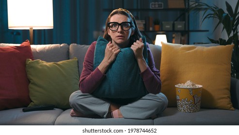 Frightened Woman Sitting On The Couch And Watching A Horror Movie On TV, She Is Hugging A Pillow