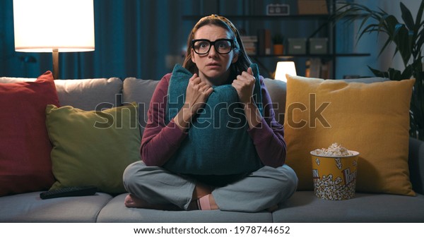 Frightened woman sitting on the couch\
and watching a horror movie on TV, she is hugging a\
pillow