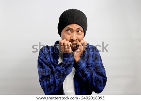Frightened young Asian man nervously bites his fingernails, gazing anxiously at the camera, visibly perturbed by something scary, experiencing a mix of anxiety, horror, and concern, white background