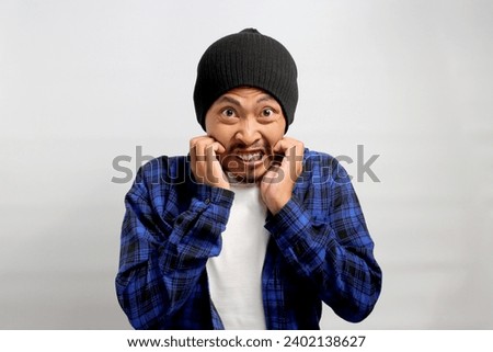 Frightened young Asian man nervously bites his fingernails, gazing anxiously at the camera, visibly perturbed by something scary, experiencing a mix of anxiety, horror, and concern, white background