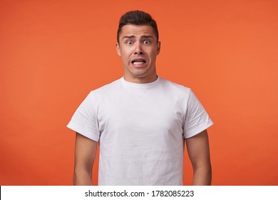 Frightened young brown-eyed brunette man with short haircut rounding amazedly his eyes while looking scaredly at camera, standing over orange background