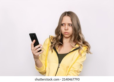 A frightened young caucasian blonde woman with wavy hair holds a mobile phone in her hand looking suspiciously at the incoming call from an unknown number isolated on a white background. Fraud